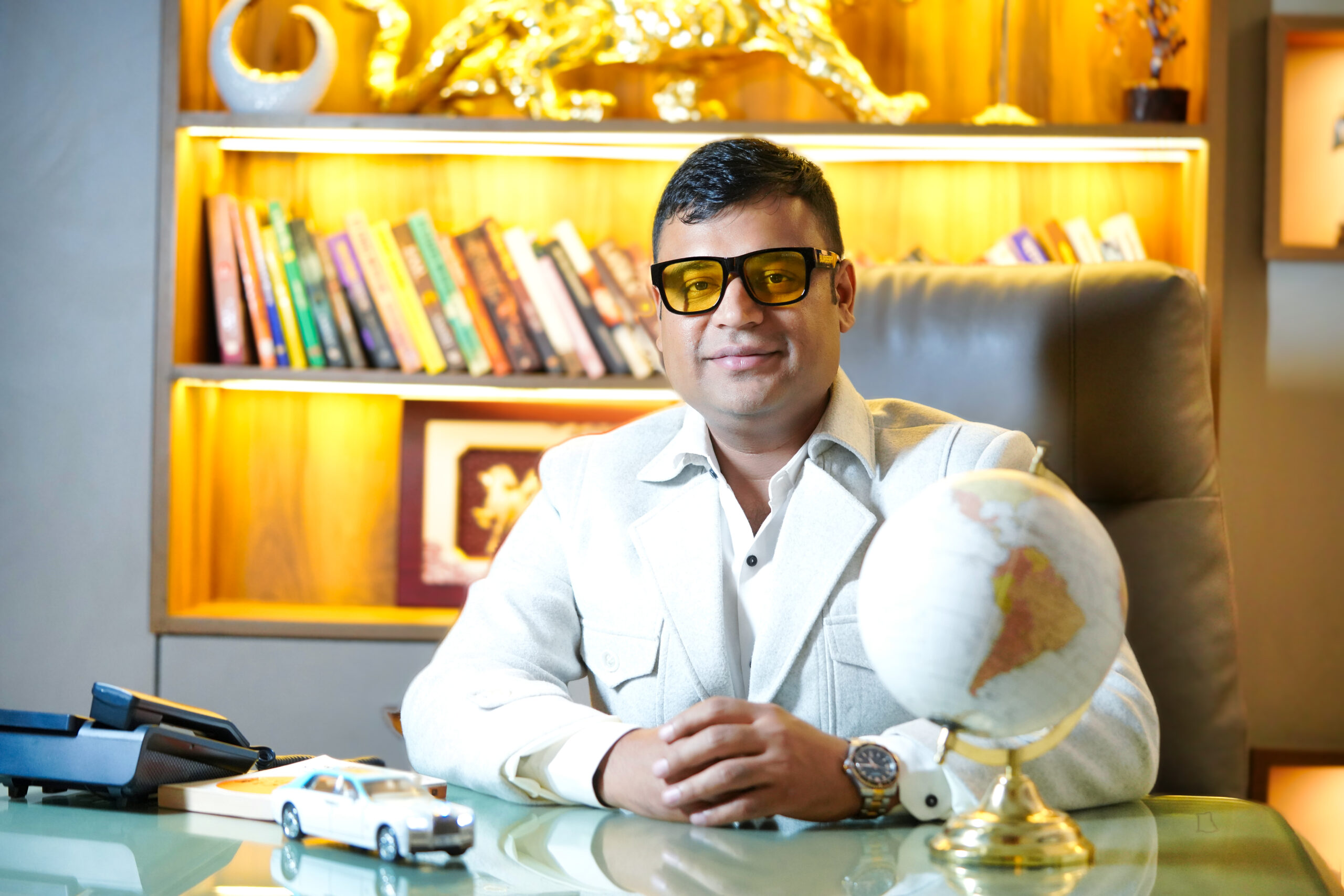 Business professional sitting at an office desk with bookshelf background, wearing formal attire and glasses, symbolizing leadership and success